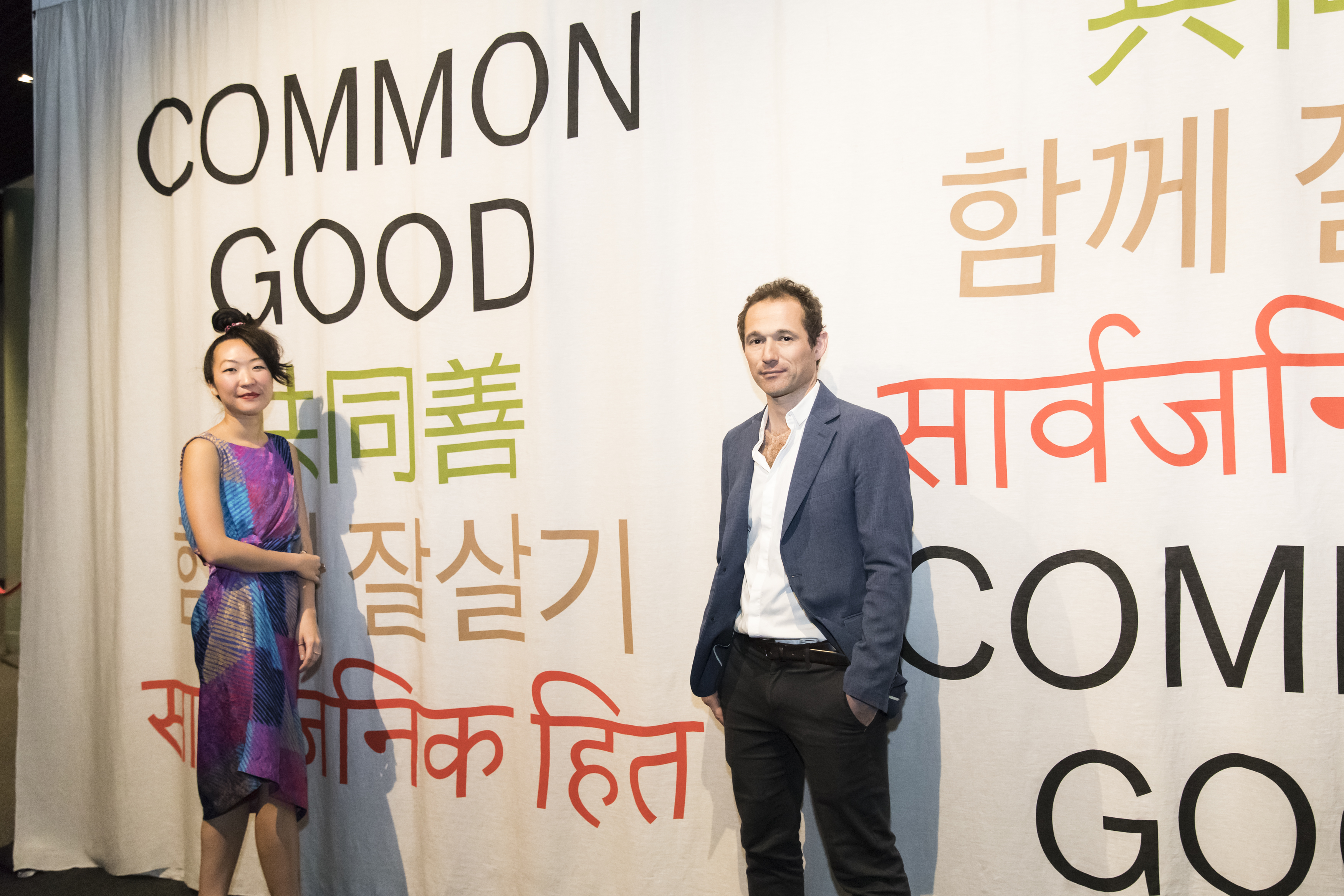 Man and woman posing in front of a curtain with Common Good written on it in various languages. 
